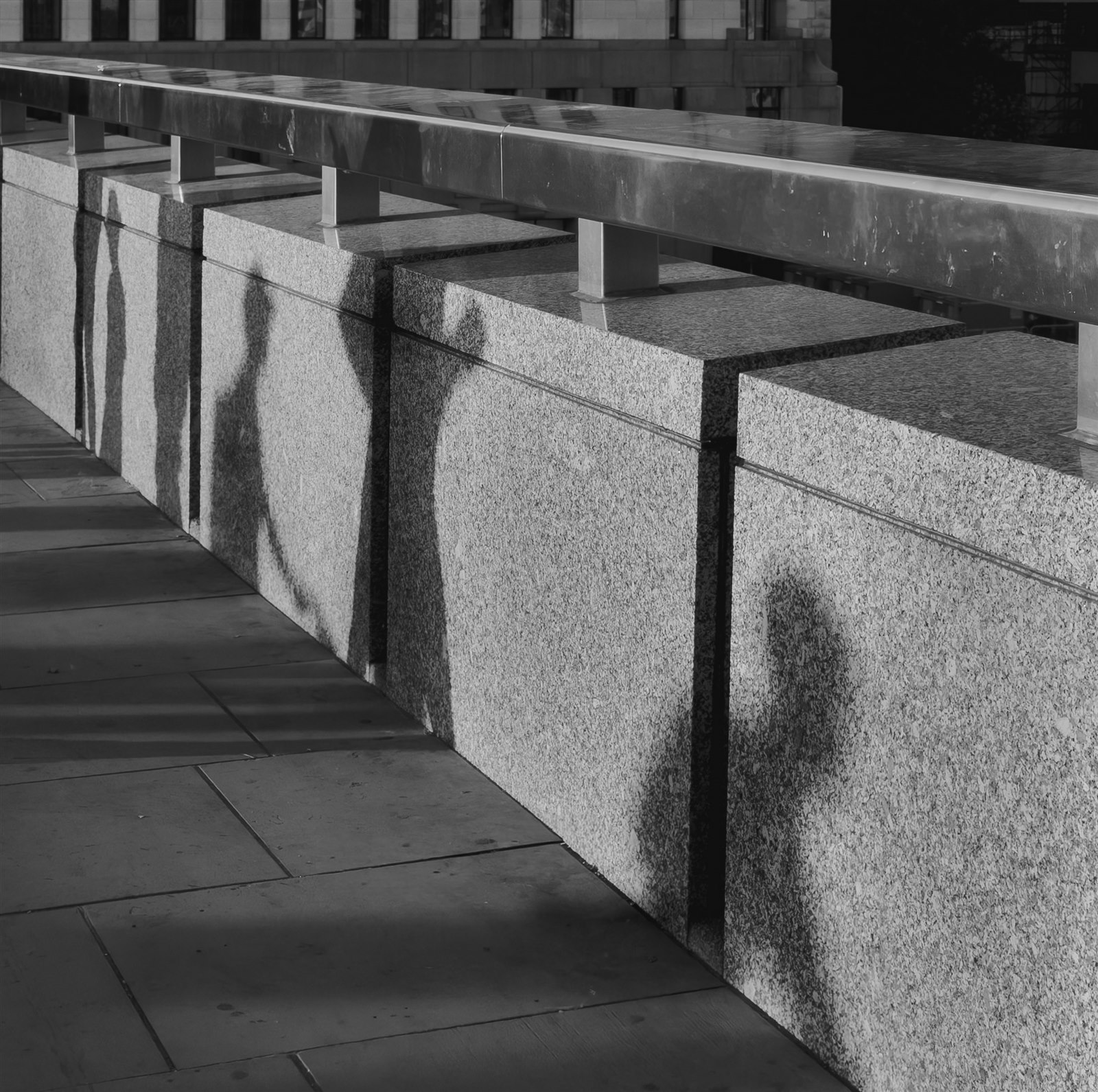 Black-and-white photograph of a human shadow falling across textured stone blocks beneath a metal railing.