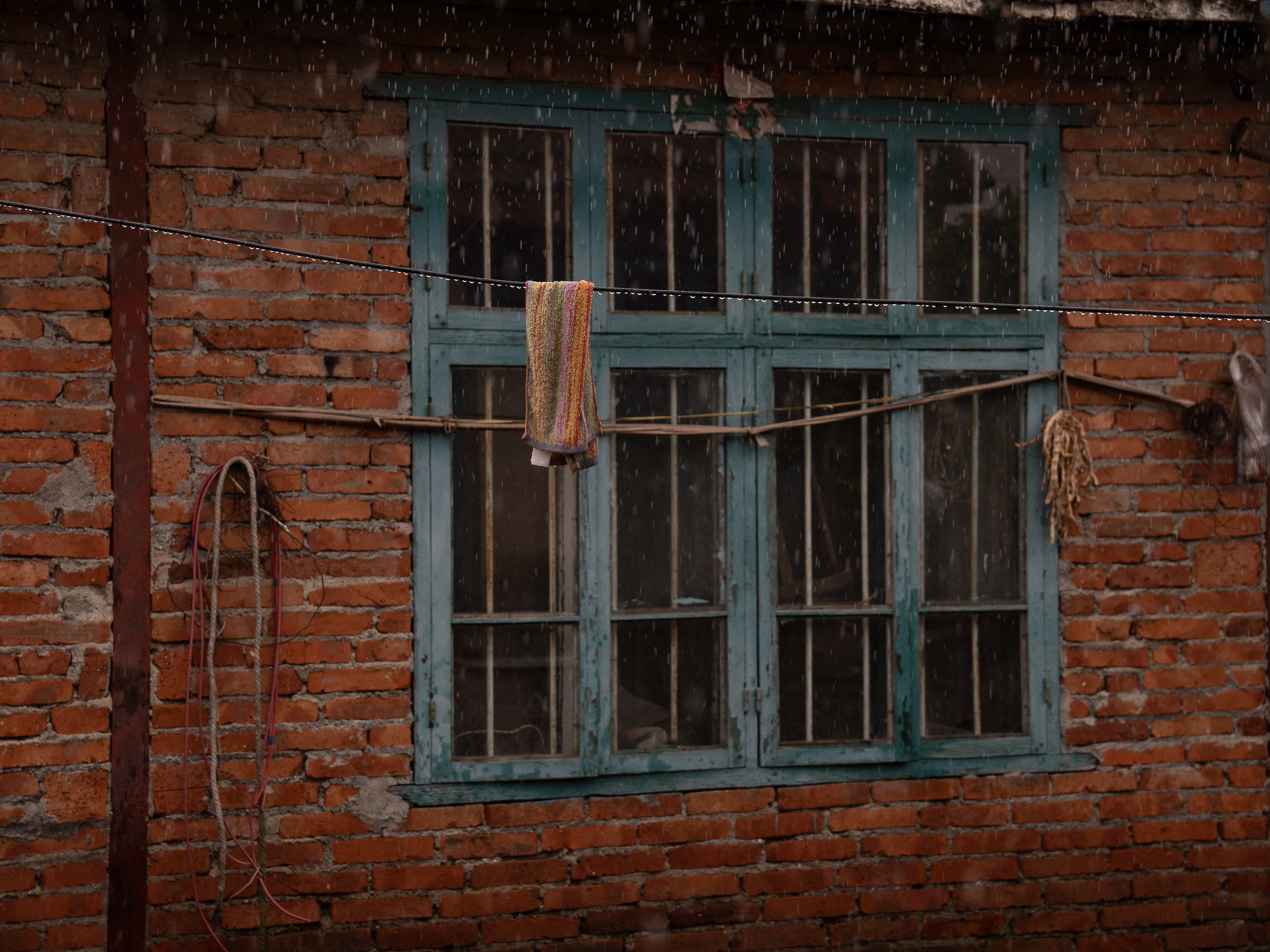 Rain falling in front of a teal wooden window in a brick wall, with a towel hanging on a line.