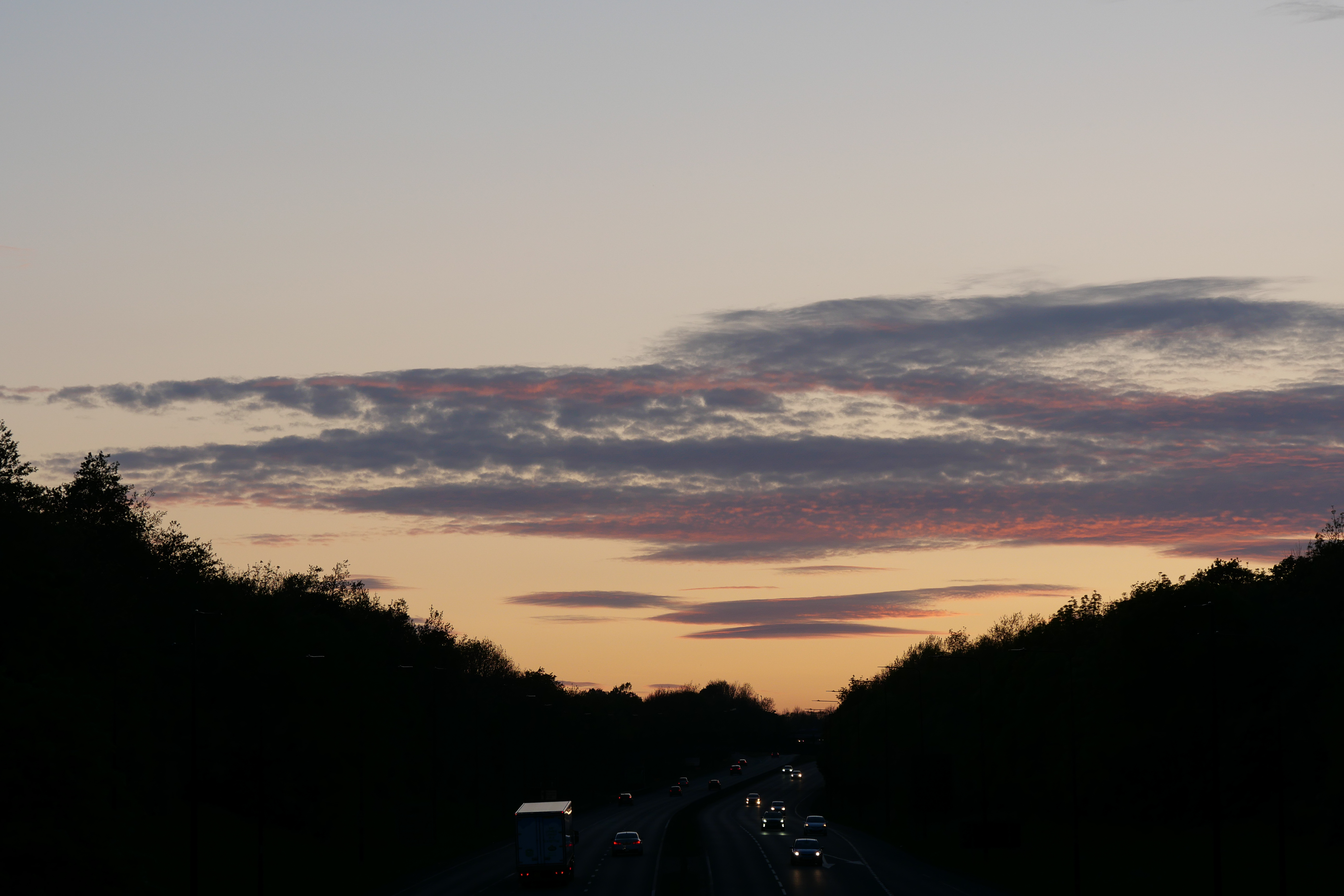Pink and violet sunset clouds above a road bordered by dark trees.